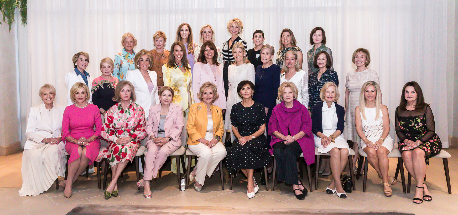A large group of women in colorful formal attire, including distinguished women in leadership, pose together for a group photo indoors, standing and sitting in several rows in front of a white curtain at the legacy luncheon.