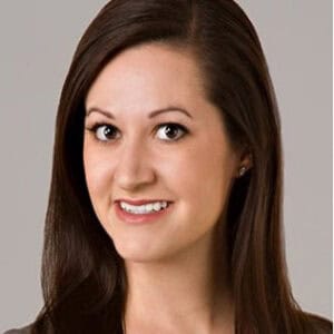 A woman with long straight brown hair and fair skin smiles at the camera. She is wearing a grey top and stud earrings, with a neutral grey background.
