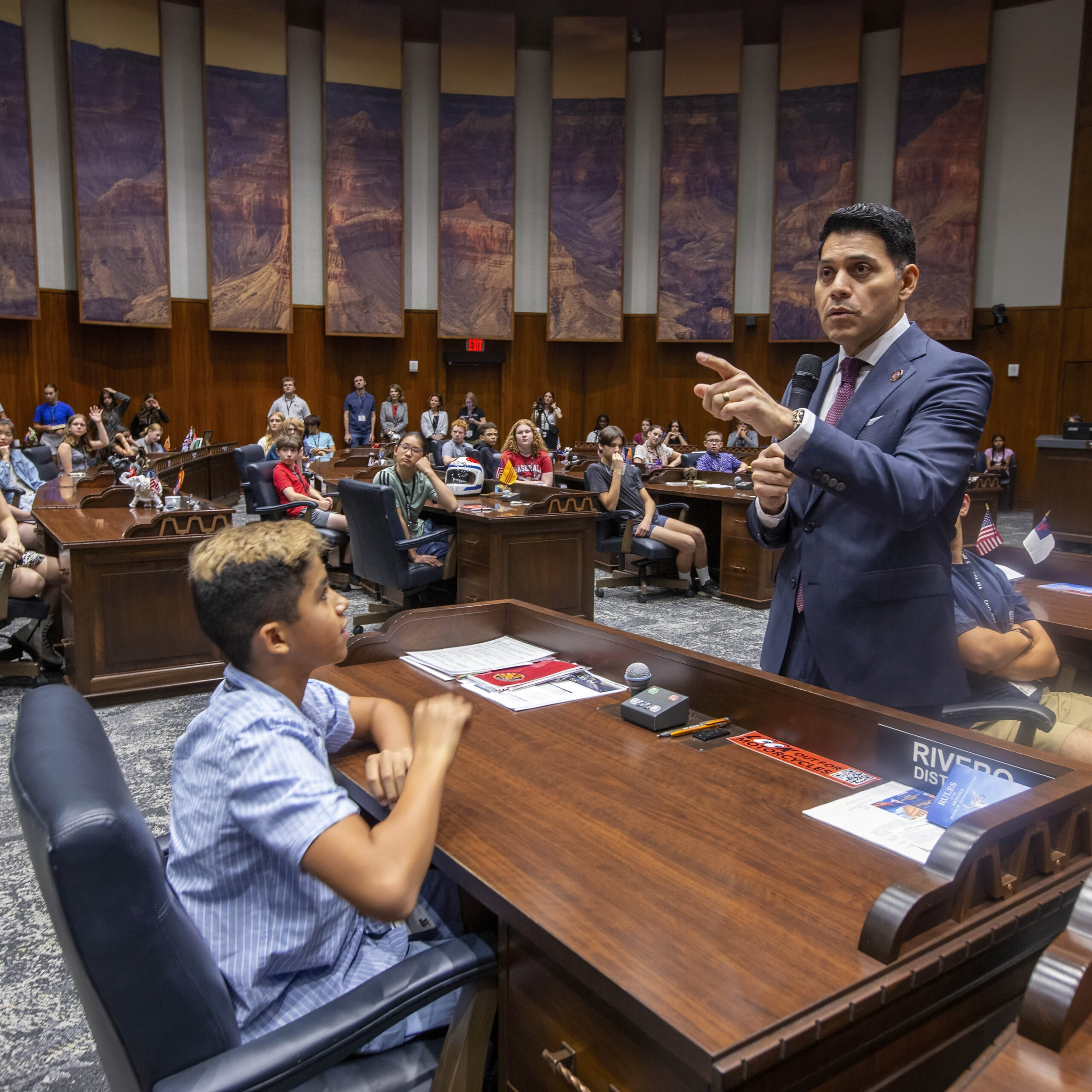 June 11, 2025 Camp SDO A man in a suit speaks to a seated boy in a government chamber filled with people. The audience listens attentively, creating a formal and educational atmosphere focused on building thoughtful citizens.