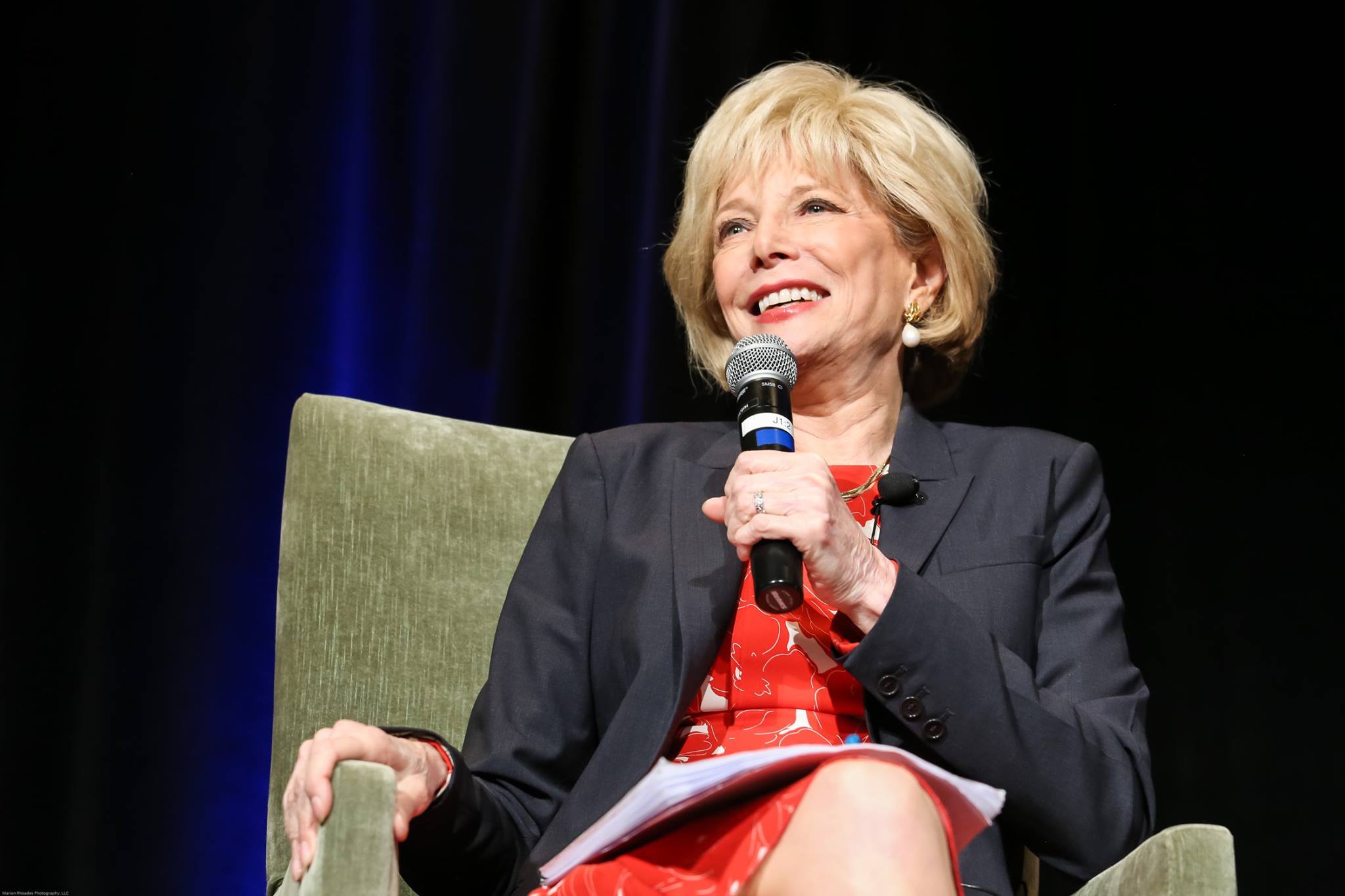 A woman with short blonde hair is sitting in a chair, smiling and holding a microphone. She is wearing a red dress with a black blazer. The background is dark, with a hint of blue light.