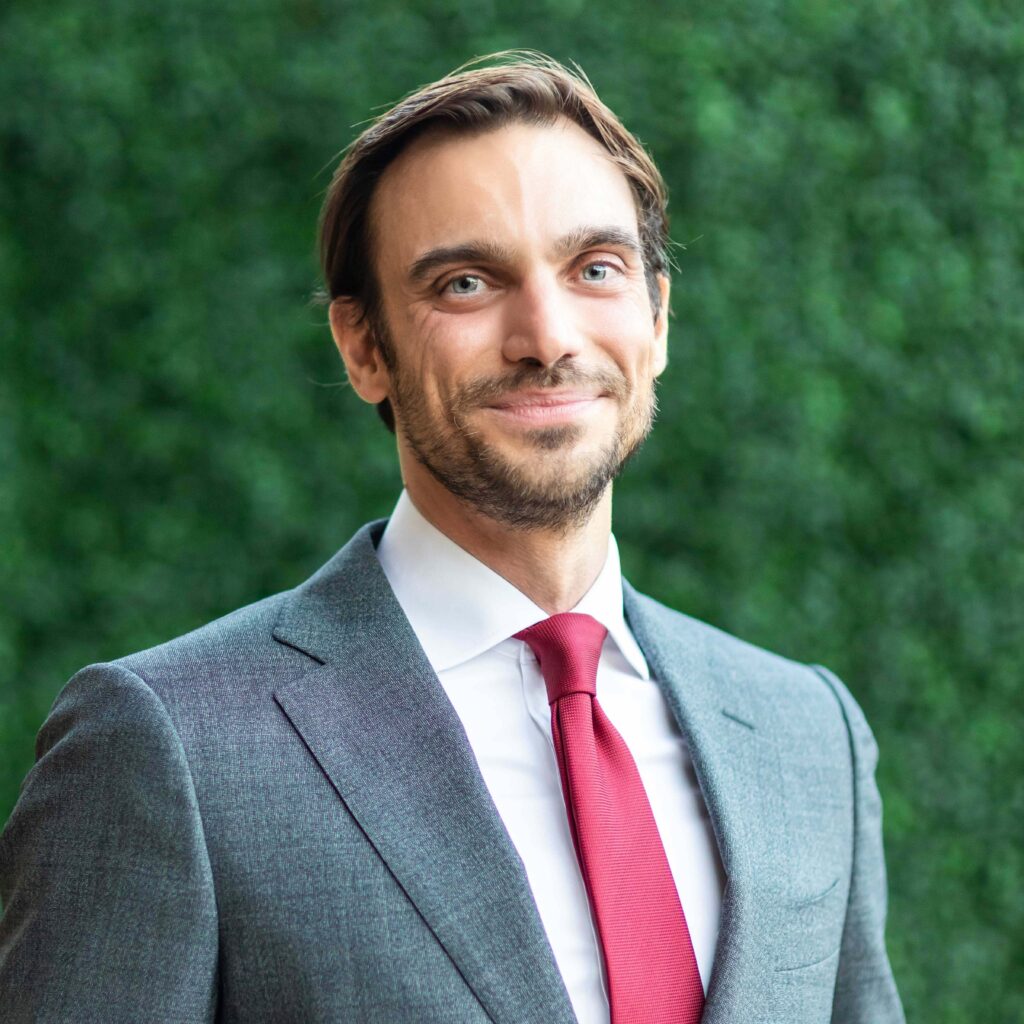 A person wearing a gray suit, white shirt, and red tie stands smiling in front of a leafy green background.