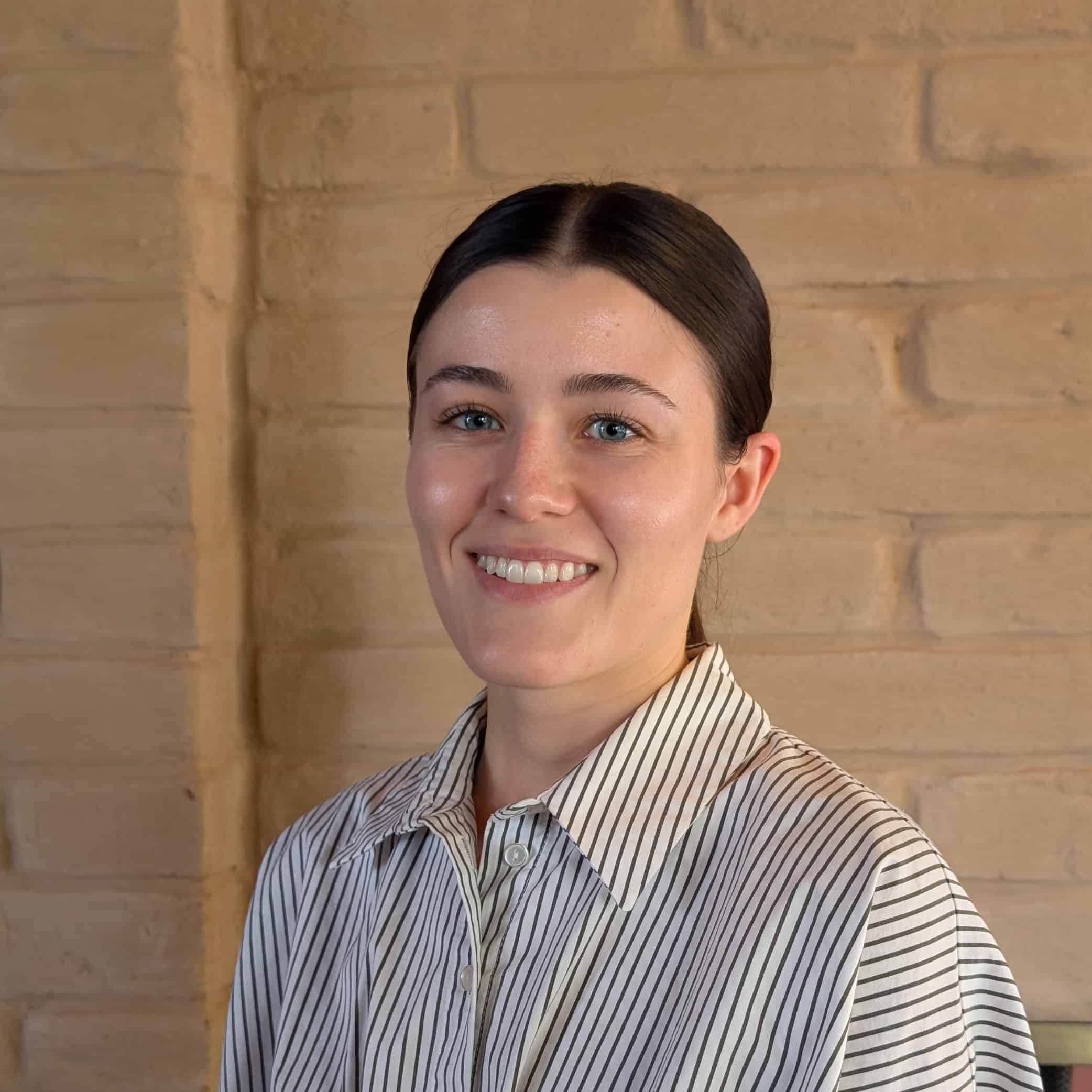 Lily Oswald, a woman with dark hair pulled back, smiles at the camera. She wears a white shirt with thin black stripes and stands in front of a tan brick wall.