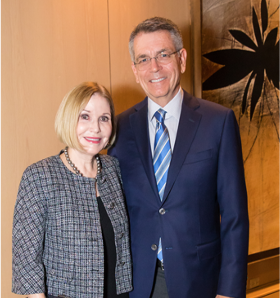 A man and a woman stand together indoors, smiling at the camera. The woman wears a checkered jacket and the man is in a suit with a blue striped tie. A large abstract artwork is visible in the background.