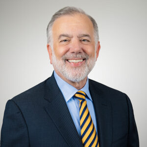 A smiling middle-aged man with gray hair and a beard is wearing a dark pinstripe suit, a light blue shirt, and a yellow and blue striped tie. The background is a light gray gradient.