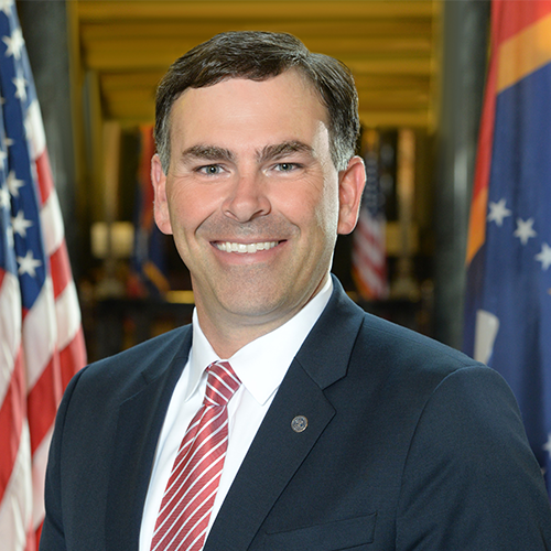 A man in a suit and red striped tie smiles at the camera. He stands between two flags, one of which is the American flag. The background appears to be an indoor setting with more flags visible.