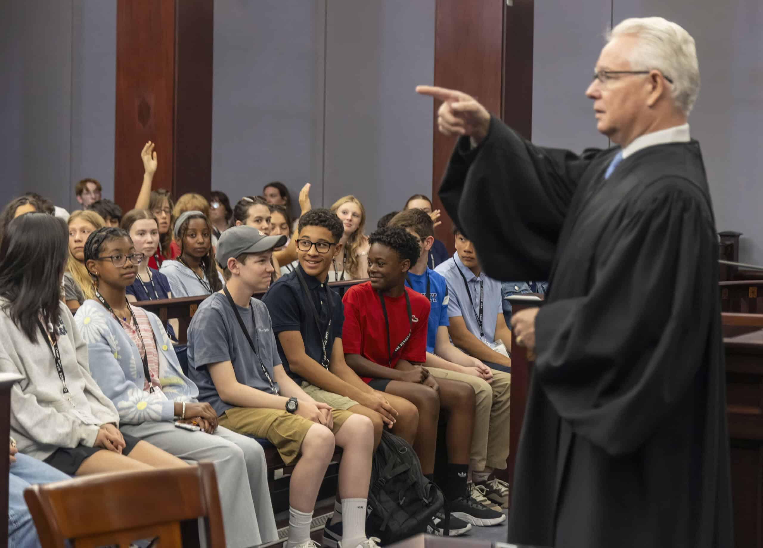 A judge in a black robe gestures while speaking to attentive students in a courtroom, some with hands raised, as The Citizen hosts an educational visit or tour.
