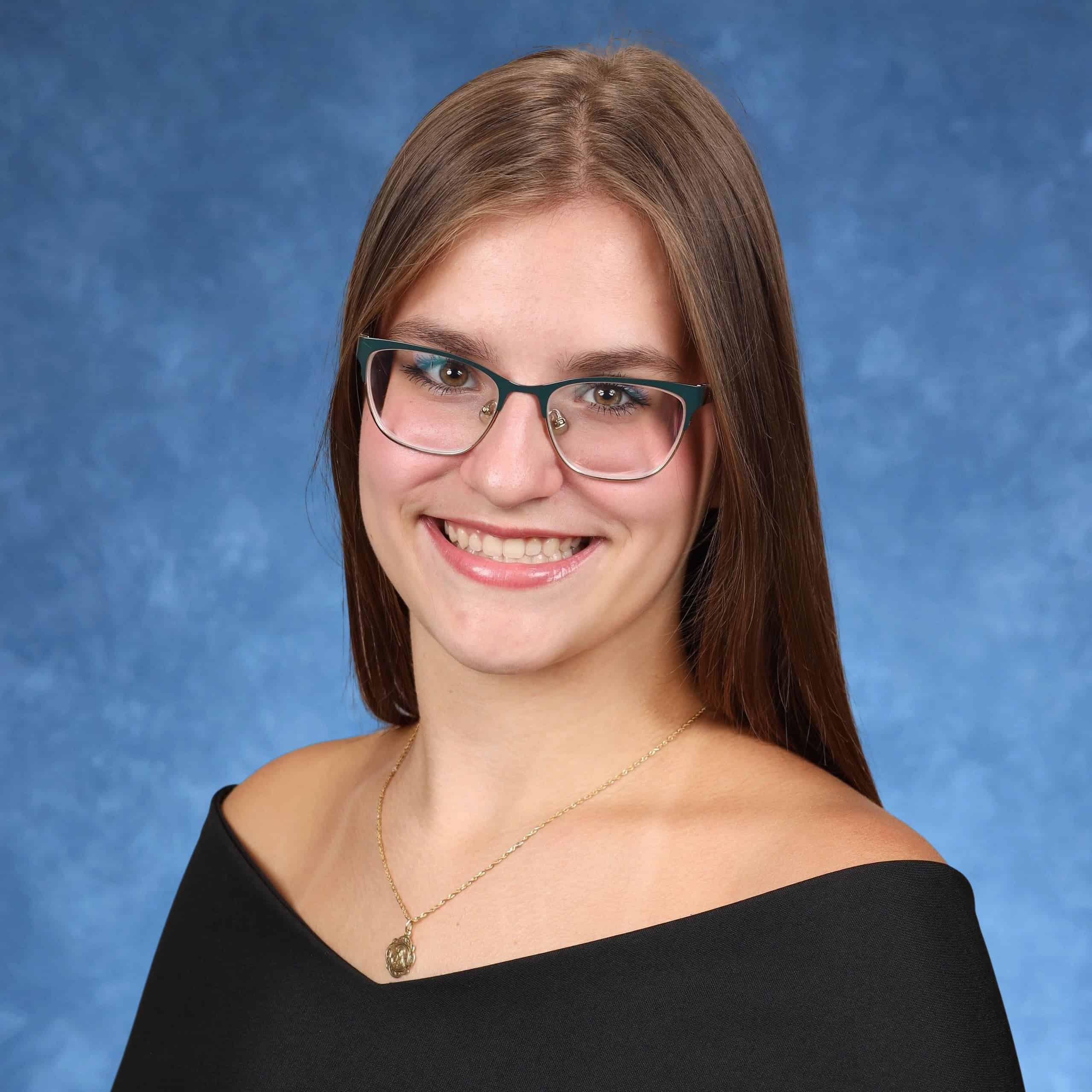 A young woman with straight brown hair, wearing glasses, a black off-shoulder top, and a gold necklace, smiles confidently in front of a blue studio background—embodying the spirit of Scholarship Winners.