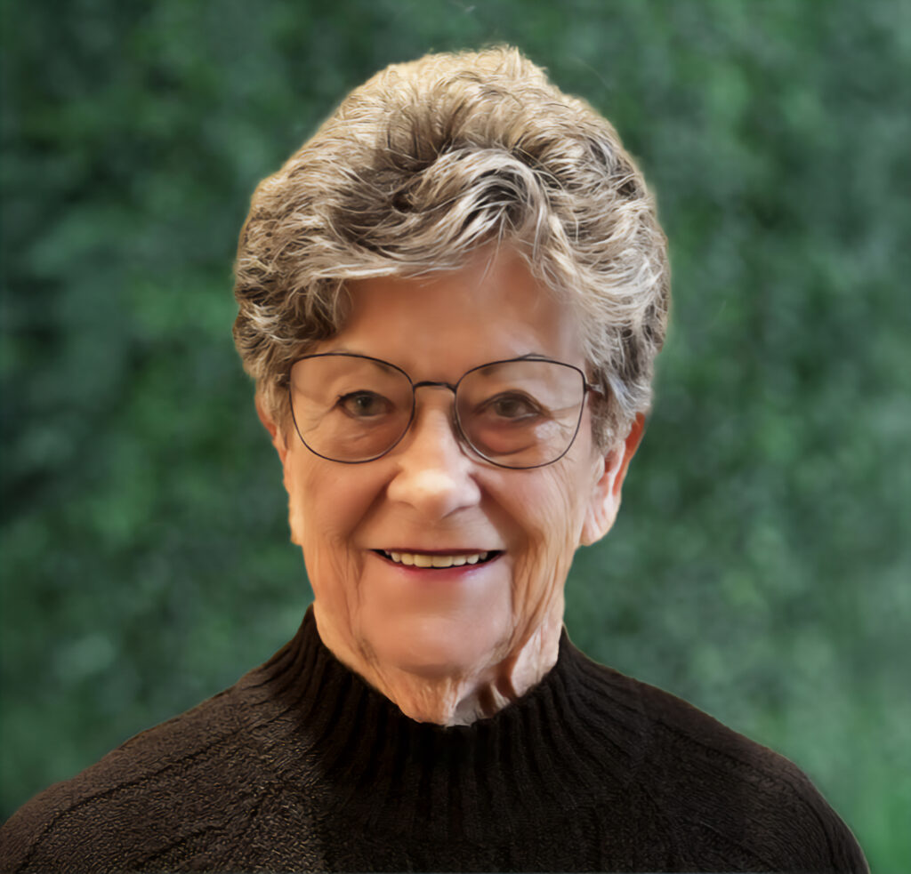 An older woman with short gray hair and glasses, wearing a black sweater, smiles at the camera. The background is a blurred green foliage.
