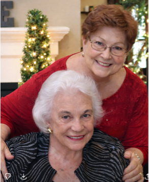 Two smiling older women pose together indoors. One, with short white hair and a zebra-striped jacket, is seated, while the other stands behind her in a red top, hugging her shoulders. A decorated tree twinkles in the background at The Citizen.