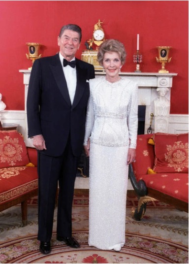 A man in a black tuxedo and a woman in a long white gown stand smiling together in an elegant red room, as if ready for a history dinner amid ornate furniture and a white fireplace.