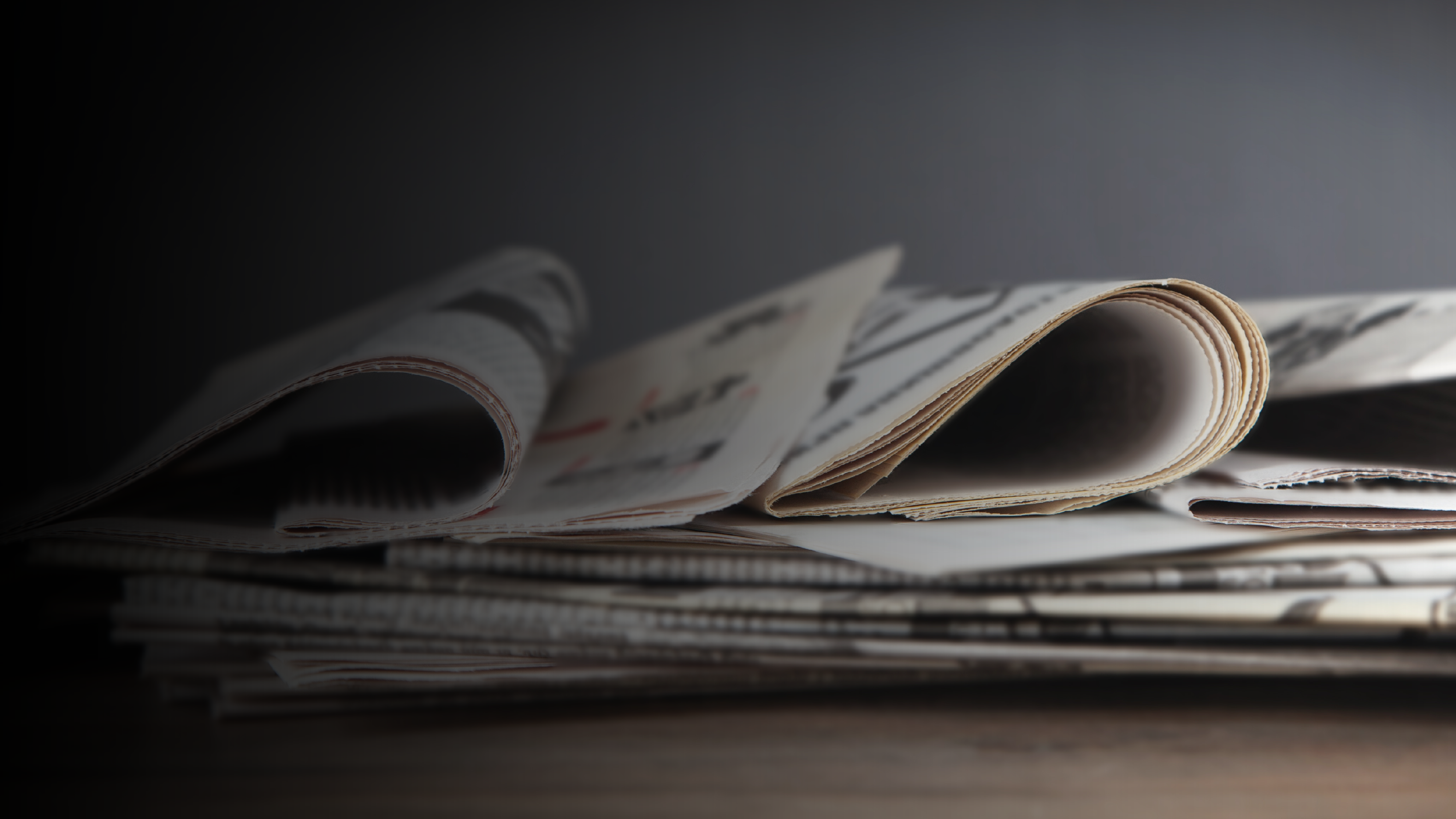 A close-up of a stack of newspapers with some pages slightly curled, lying on a wooden surface against a dark, blurred background—one headline mentions Sandra Day O'Connor.
