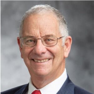 Smiling older man with short gray hair, glasses, and a suit with a red tie, posed in front of a gray blurred background.