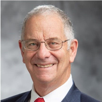 Smiling older man with short gray hair, glasses, and a suit with a red tie, posed in front of a gray blurred background.