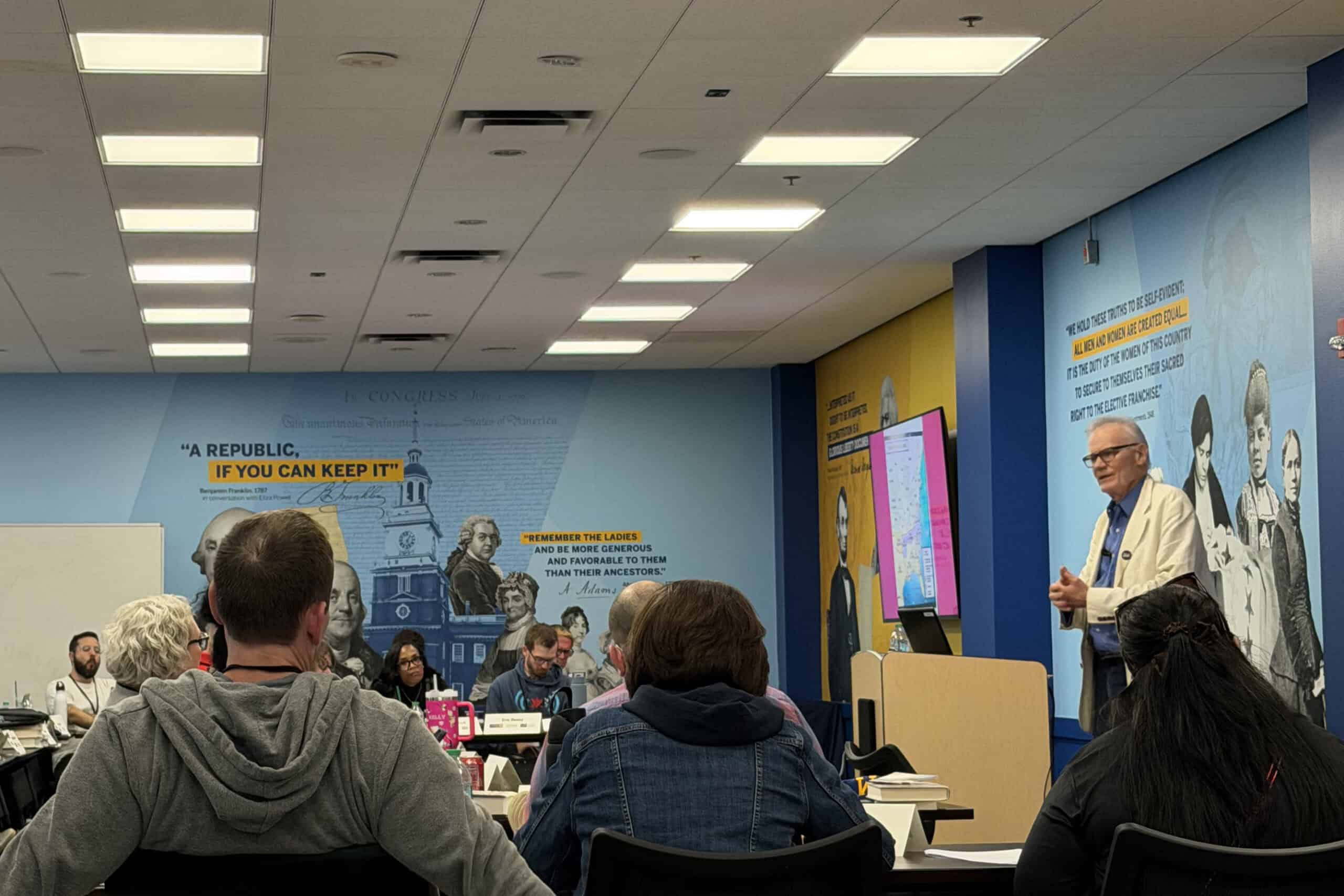 A classroom setting with a lecturer at a podium addresses seated students. The walls display historical quotes and images, including one reading, “A REPUBLIC, IF YOU CAN KEEP IT,” inspiring every Citizen in the room.