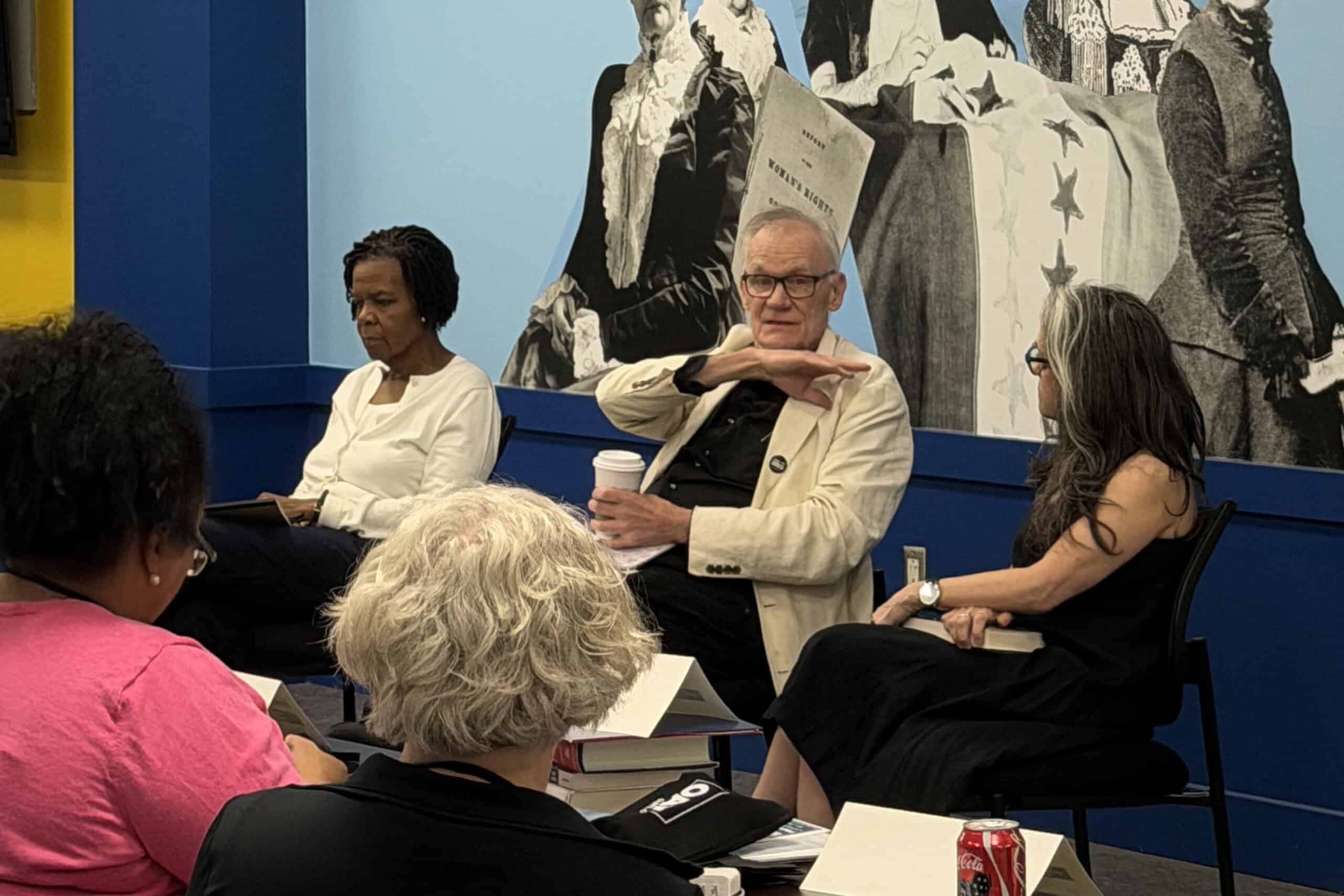 Three people sit in front of an audience, engaged in a discussion for The Citizen. One man gestures while holding a cup, flanked by two women. Behind them is a large historical mural, with books and a soda can on the table.