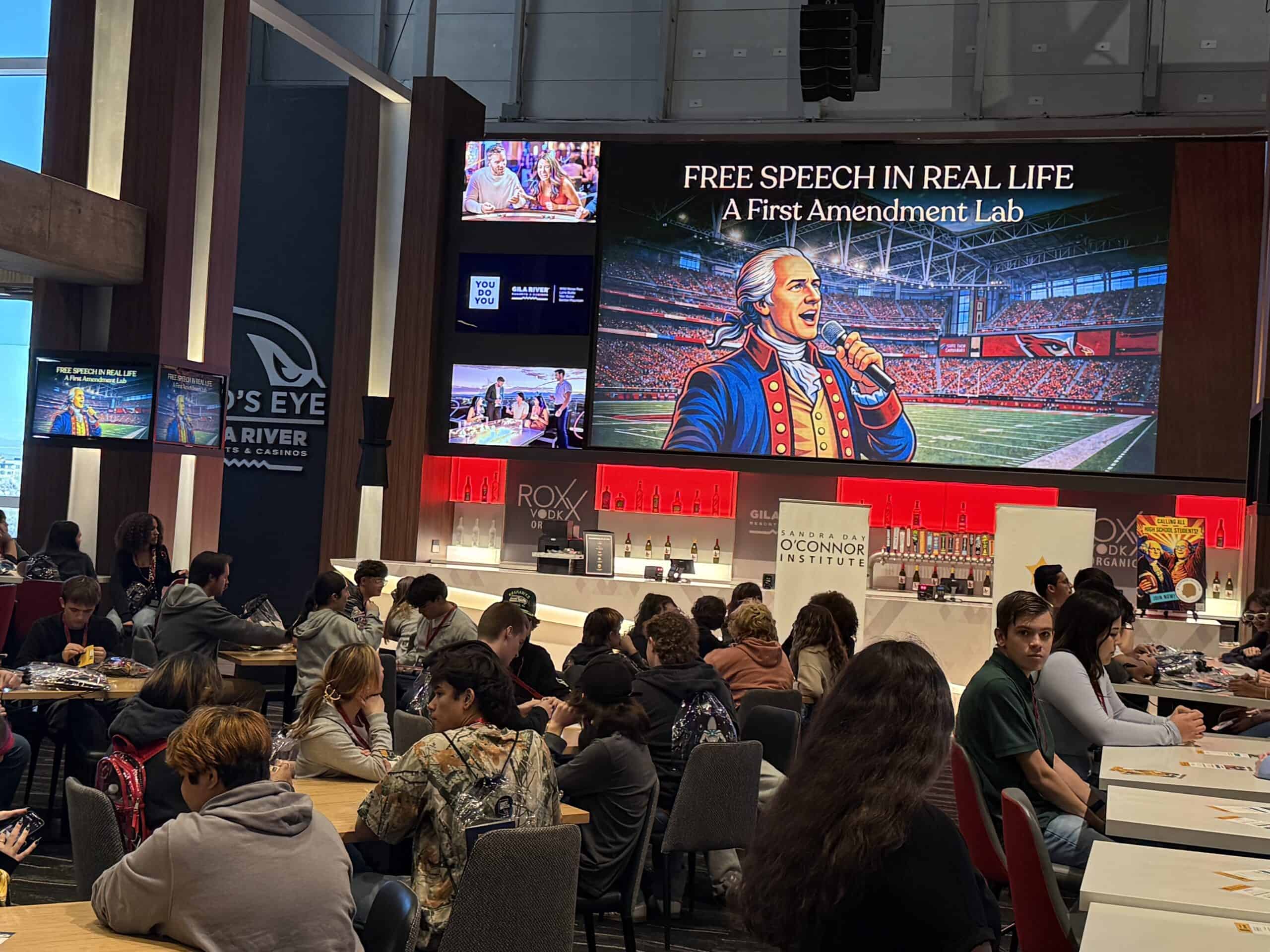 A large group sits at tables in a modern hall, listening to a presentation. Onstage, a screen displays “FREE SPEECH IN REAL LIFE: A First Amendment Lab,” marking America’s 250th Anniversary with an illustrated historical figure speaking.