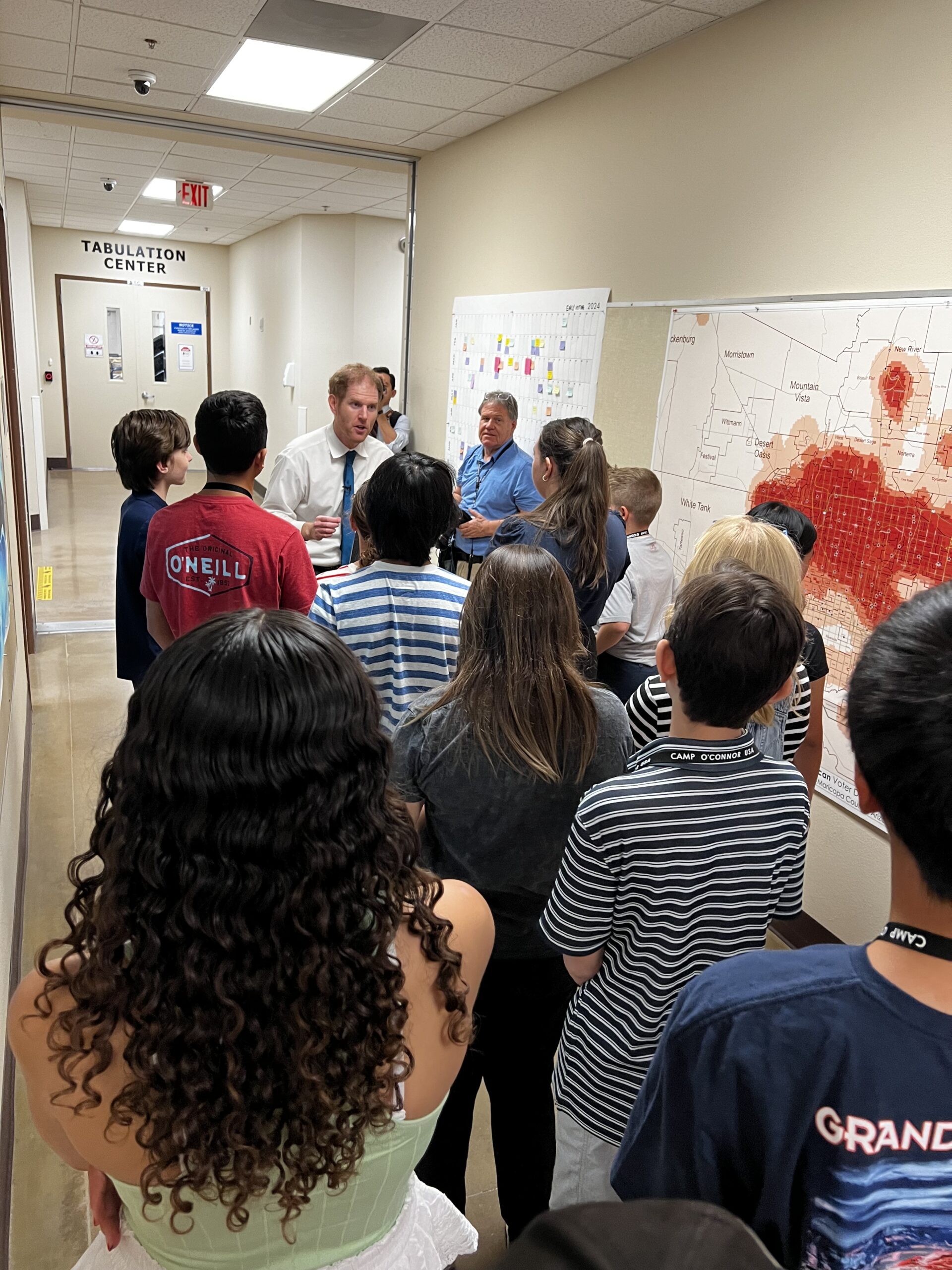 A group of people gather in a hallway near a wall with maps and charts. A man in a white shirt appears to be speaking to the group. The setting resembles an office or an educational environment.