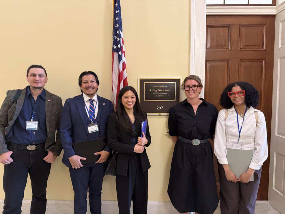 Five people stand smiling in front of an office door with a sign reading “Rep. Greg Stanton, Arizona, 207,” showcasing their Capital Connections. An American flag is next to the group, all dressed in business or professional attire.