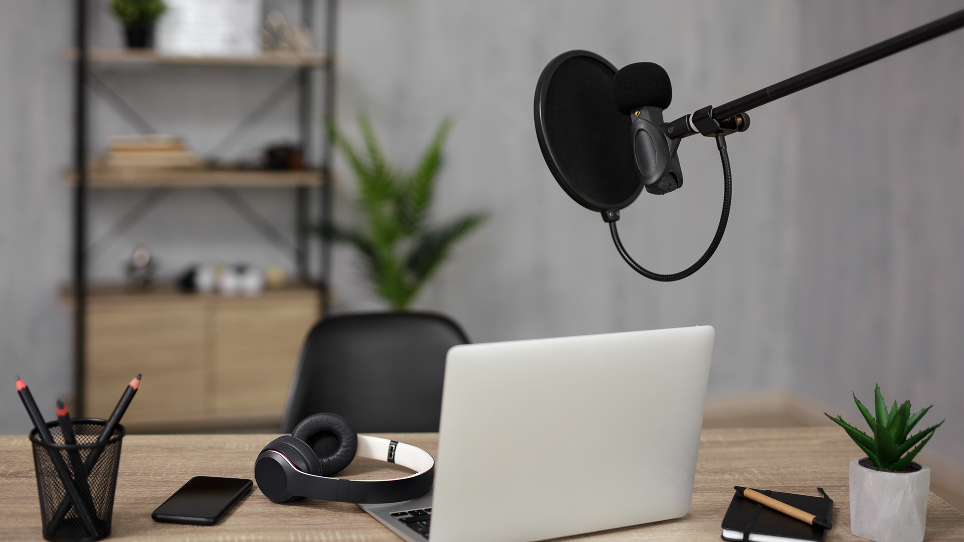 A modern podcasting setup featuring a laptop, microphone with pop filter, and headphones on a wooden desk. A pencil holder, notebook, and small potted plant decorate the workspace. A shelf and plant are visible in the background.