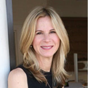 A woman with long blonde hair and a black sleeveless top smiles while standing indoors near a white wall.
