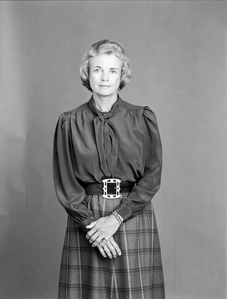 A citizen stands against a plain background: a woman with short, light hair, wearing a long-sleeve blouse with a bow and a belted plaid skirt. Her hands are clasped in front of her, embodying the quiet poise of The Citizen.