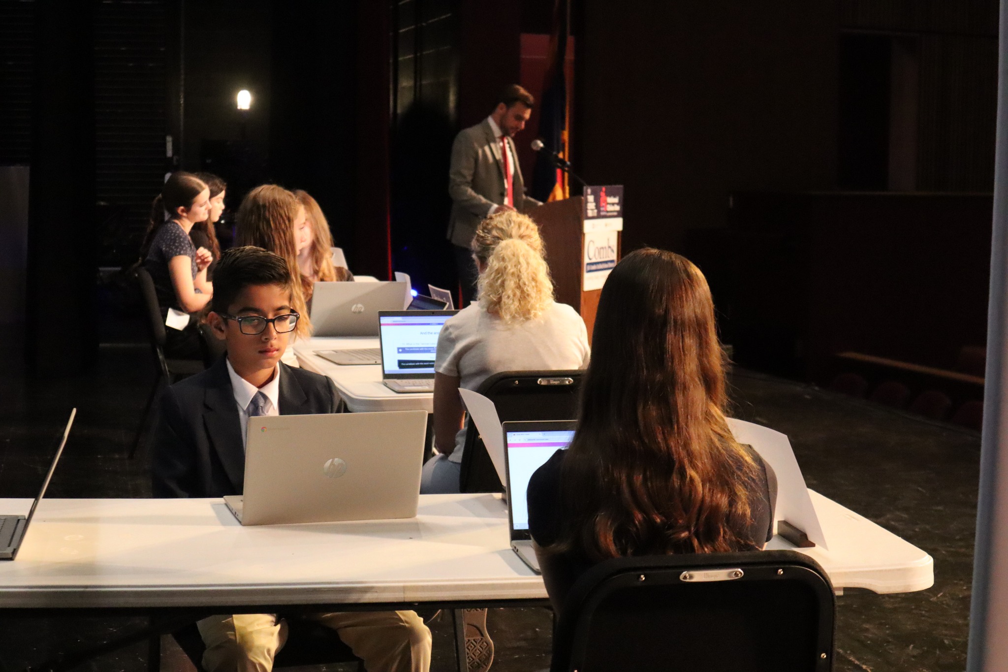 QueenCreekCivicBee Several students in formal attire sit at tables with laptops on a stage, facing away from the audience. A man stands at a podium in the background, speaking into a microphone.