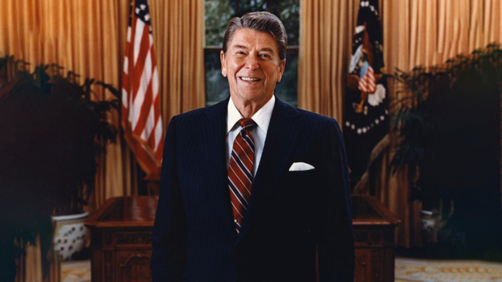 A man in a dark suit and striped tie stands smiling in front of a desk, as if ready for a history dinner, with an American flag and gold curtains in the background.