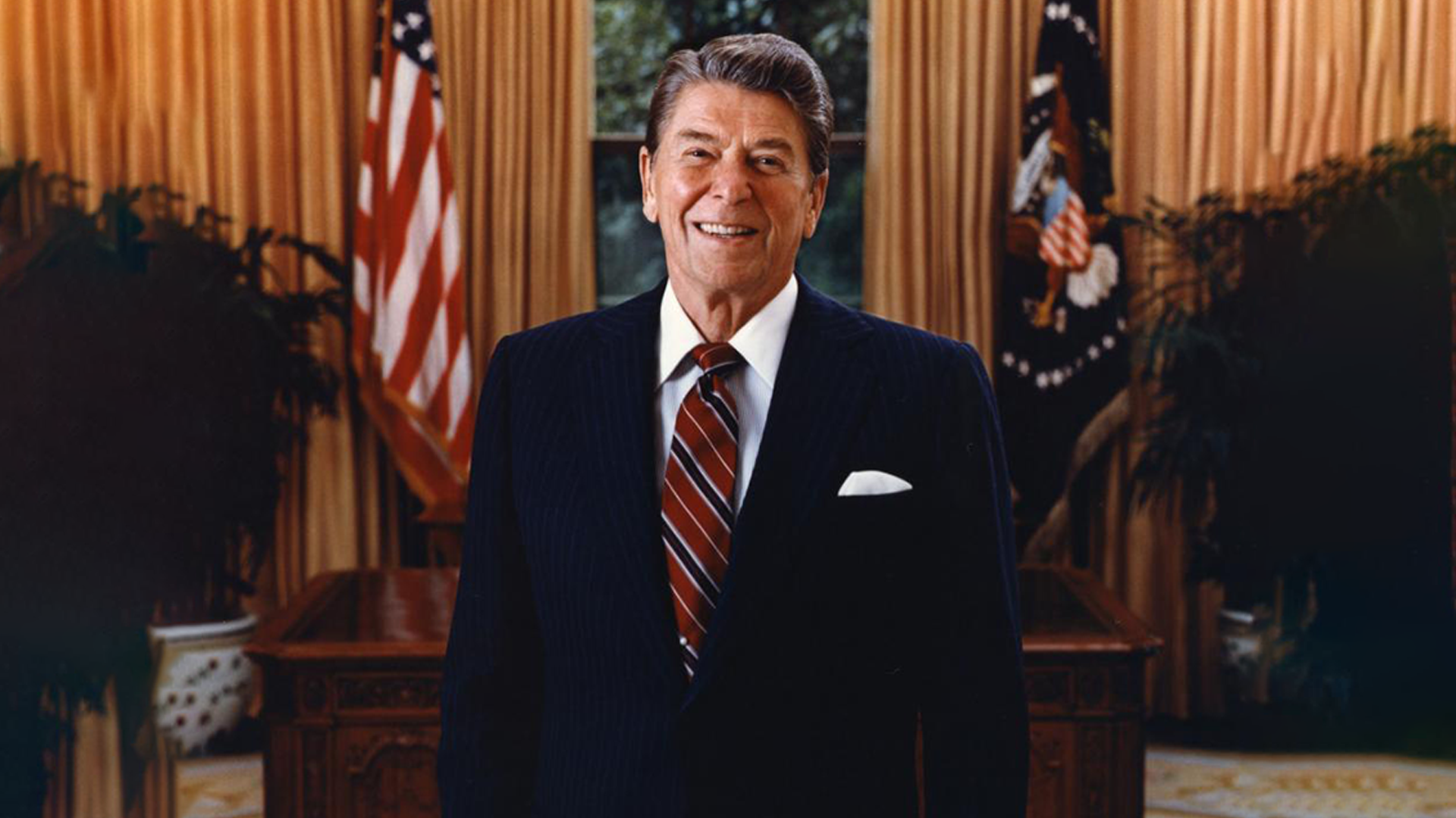A man in a dark suit and striped tie stands smiling in front of a desk, as if ready for a history dinner, with an American flag and gold curtains in the background.