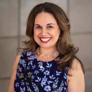 A woman with long brown hair is smiling at the camera. She is wearing a navy blue dress with a floral pattern and a pearl necklace. The background is a neutral-colored wall.