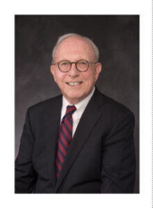 An older man in a black suit, white shirt, and red-striped tie is smiling against a dark background. He has short gray hair and is wearing round glasses.