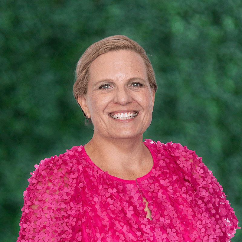 A person with short hair smiles at the camera, wearing a bright pink top with textured floral patterns. The background is a blurred green, resembling foliage.