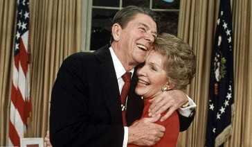 An older man in a suit and an older woman in a red outfit embrace and smile joyfully in a formal room with gold curtains and American flags, capturing the warmth of a history dinner celebration.