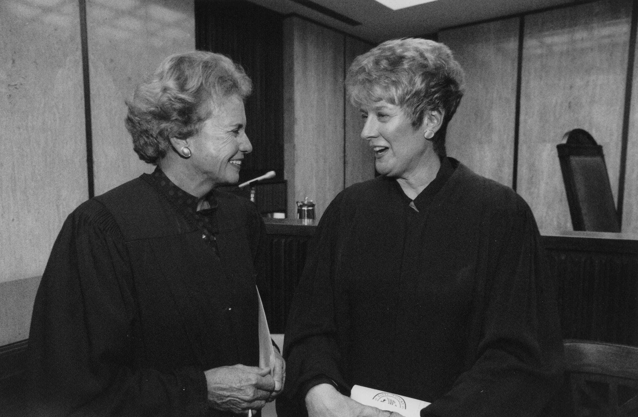 Two women in judicial robes stand in a courtroom, smiling and talking to each other. One holds a document, perhaps an important brief from The Citizen. A judge’s bench and chair are visible in the background.