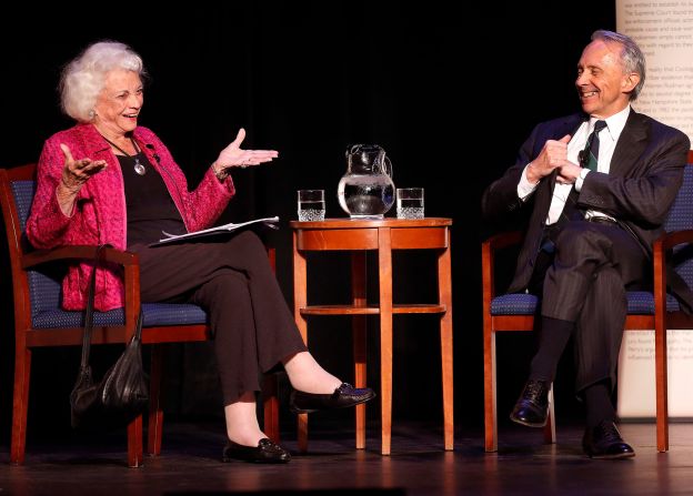 An older woman and a man in formal attire sit on stage, smiling and talking. Seated with water between them against a dark background, they engage in civil discourse, highlighting the importance of respectful conversation.