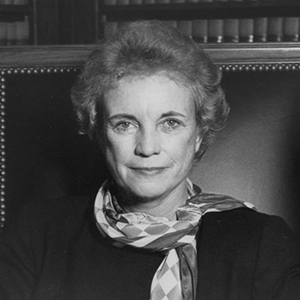 Black and white photo of a person with short, curly hair, wearing a scarf and dark top, sitting against a leather chair with nailhead trim. Books are visible in the background on a wooden shelf.