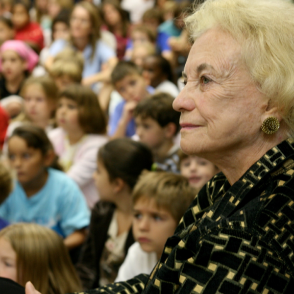SDOCamp An older woman with short blonde hair wearing a patterned jacket sits among a crowd of children. The children are seated and appear to be watching a presentation or event. The focus is on the woman's profile and attentive expression.