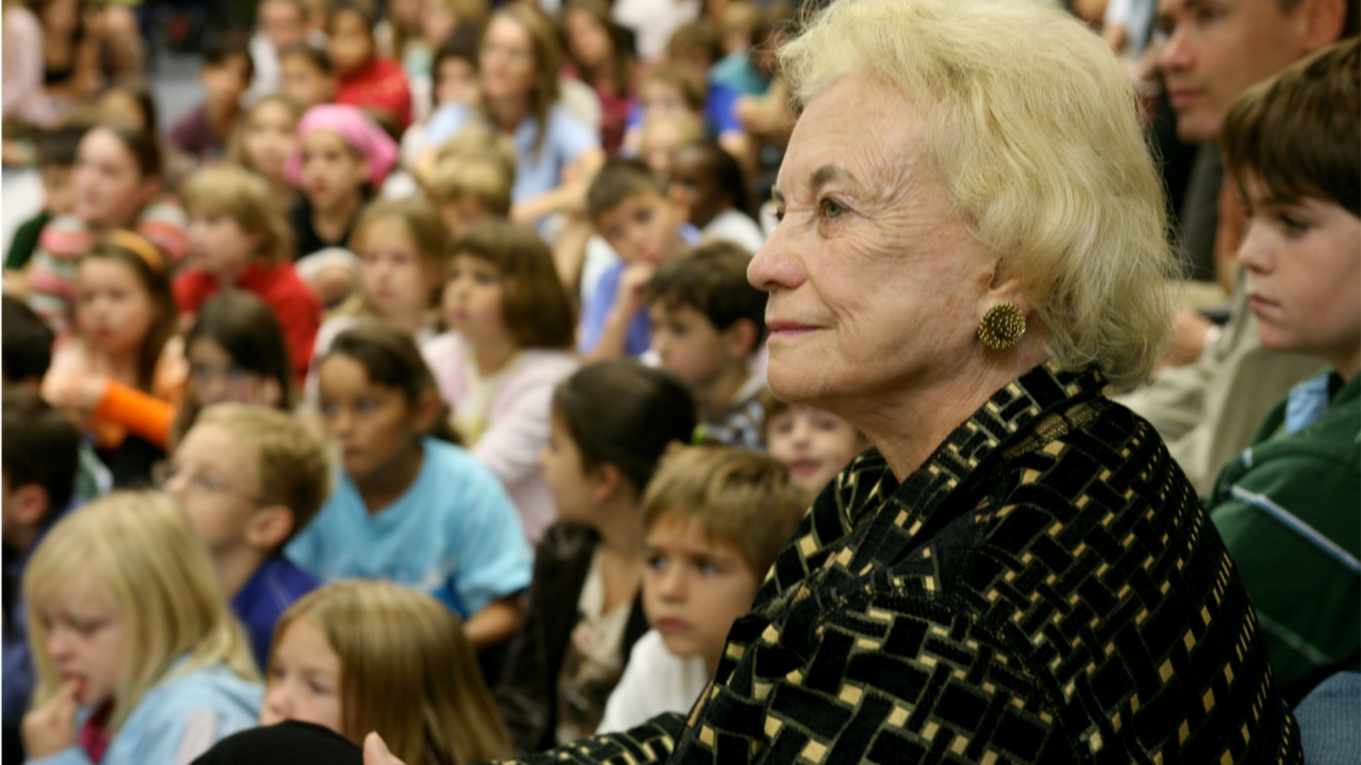 An older woman with short blonde hair wearing a patterned jacket sits among a crowd of children. The children are seated and appear to be watching a presentation or event. The focus is on the woman's profile and attentive expression.