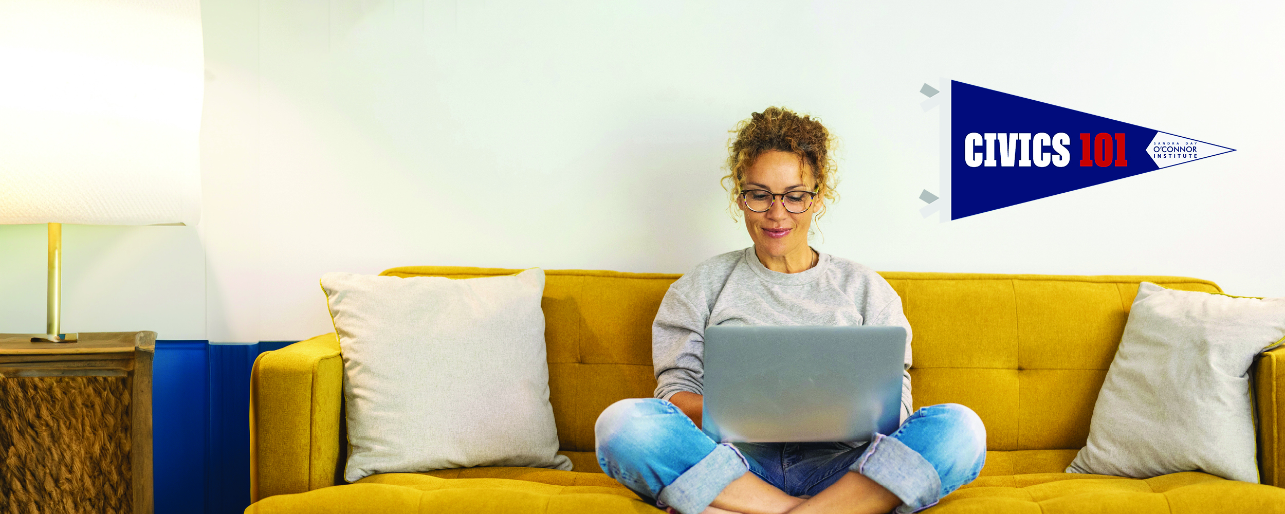 A person with curly hair and glasses sits on a yellow sofa, using a laptop. Two pillows are beside them. A lamp is on the wooden side table, and a wall flag reads "Civics 101.