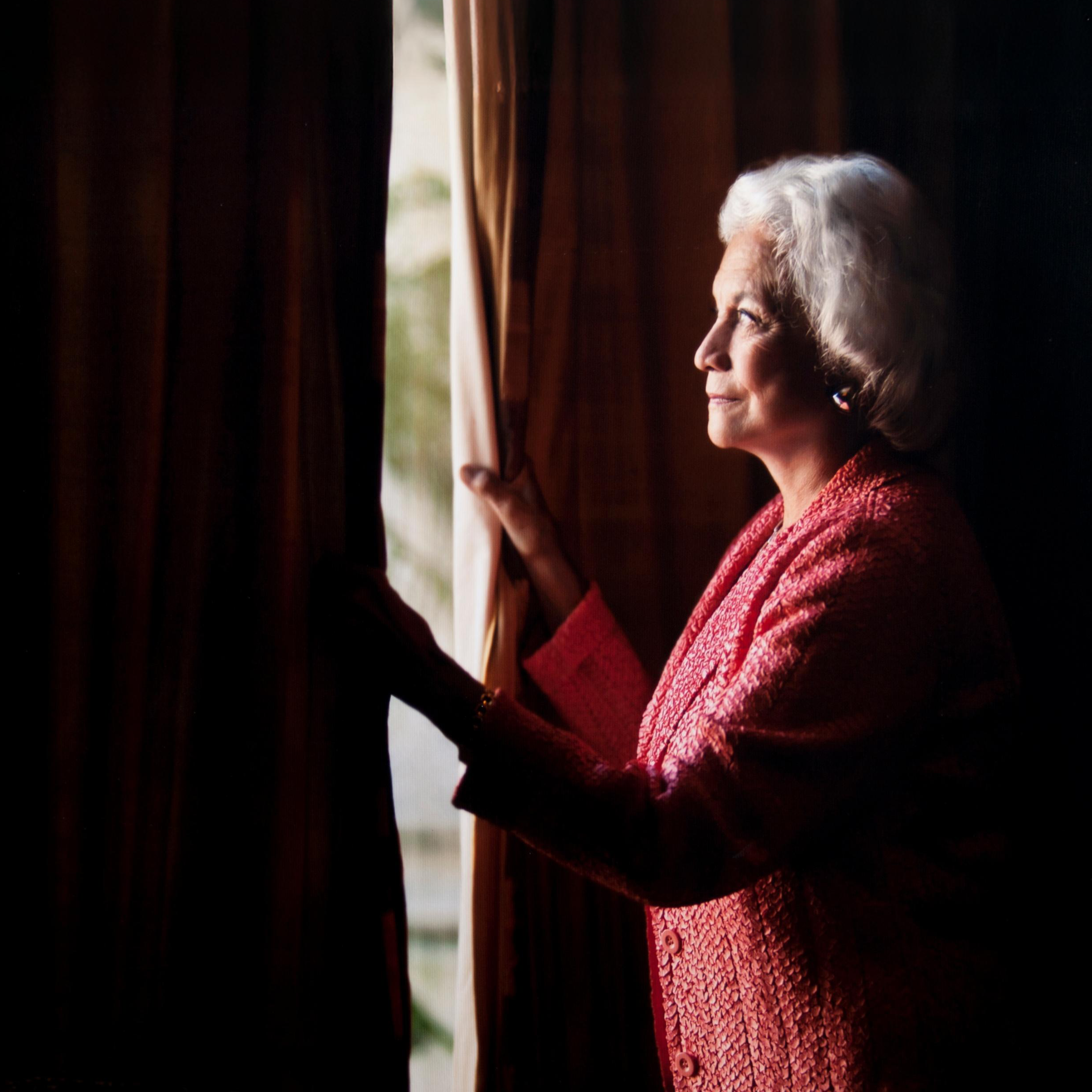 An elderly woman with short white hair stands by a window, holding the curtains open slightly, gazing outside. She is wearing a textured pink jacket. The light creates a soft glow around her.