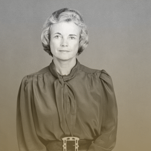 A woman with short, wavy gray hair wears a long-sleeved blouse with a necktie and a wide belt, facing the camera with a neutral expression. The simple, gradient background evokes the professional setting of Youth Programs.