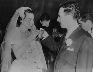 A black and white photo of a bride and groom sharing a toast. The bride, wearing a veil, smiles while holding a cup. The groom, in a suit with a boutonniere, faces her. They are surrounded by other people at the event.