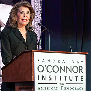 A person in a black suit stands behind a podium with a microphone, speaking at an event. The podium has a sign that reads "Sandra Day O'Connor Institute for American Democracy.
