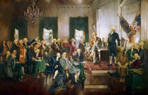 A historical painting of the Founding Fathers in a grand room signing the U.S. Constitution, with men in period clothing gathered around a table and an American flag displayed—perfectly capturing the spirit of America's 250th celebration.