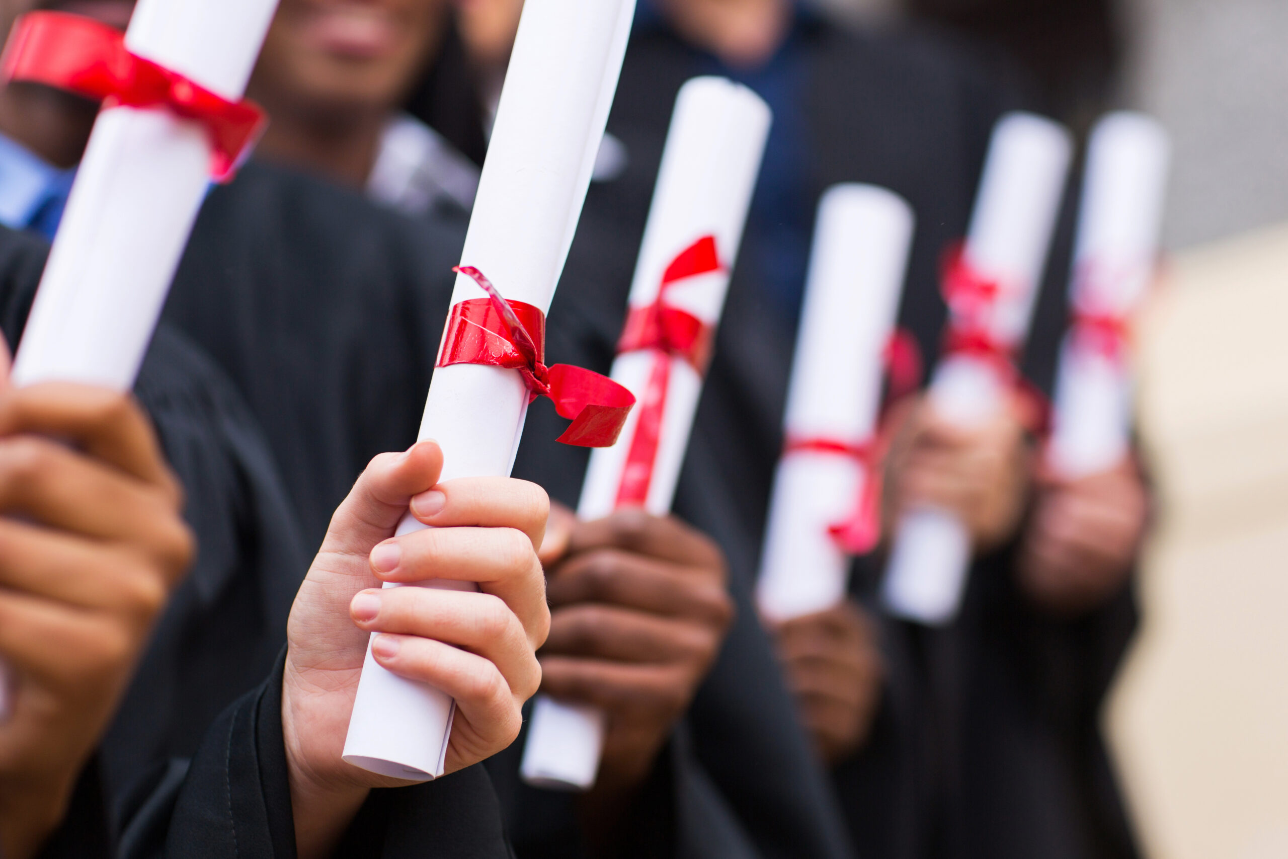 Close-up of hands holding rolled diplomas tied with red ribbons. The individuals are dressed in black graduation gowns, celebrating an academic achievement.