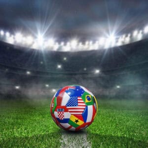 A soccer ball covered in various national flags sits on a grass field under bright stadium lights in a large, foggy arena, capturing the excitement of the World Cup.