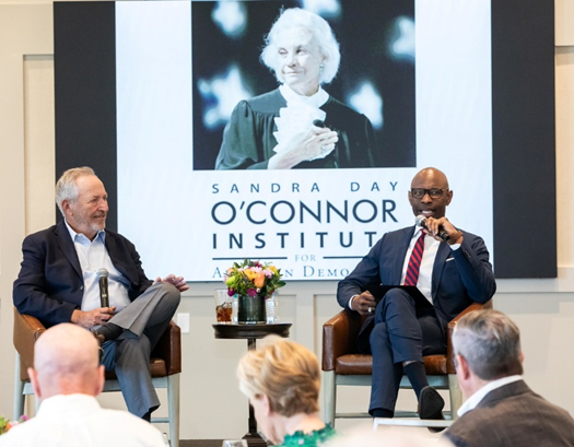 Summers1 Two men, including Dr. Summers, sit on stage with microphones, addressing an audience. Behind them, a large screen displays a photo of Sandra Day O’Connor in judicial robes and the text “Sandra Day O’Connor Institute for American Democracy.”.
