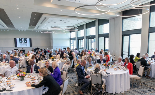 Summers2 A large group, including Dr. Summers, sits at round tables in a spacious, modern banquet hall with big windows, eating and conversing during a formal event. Flowers and plates of food are on the tables.