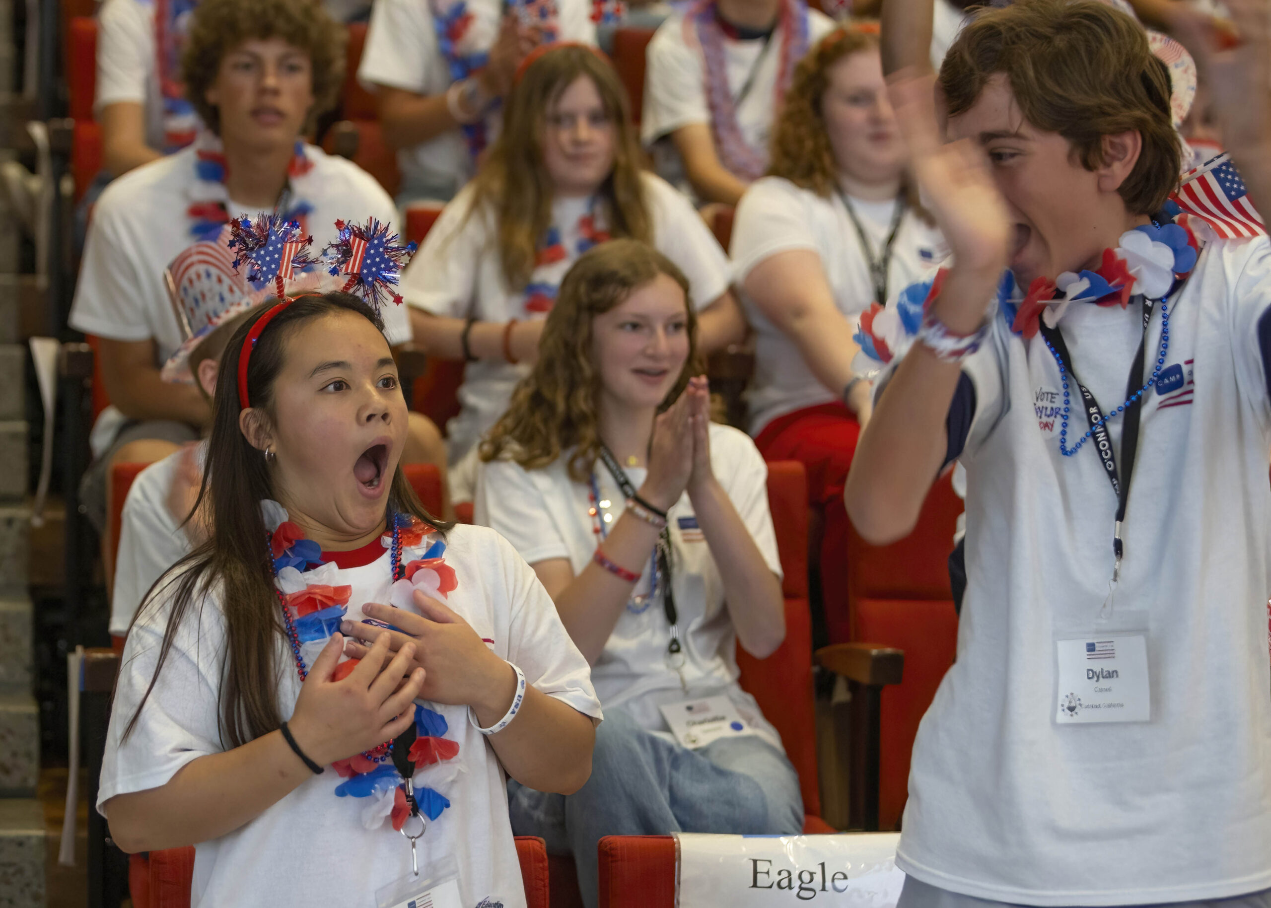June 13, 2025 Camp SDO A girl and boy wearing patriotic accessories react with excitement in an auditorium, surrounded by fellow citizens—students in matching white shirts and red, white, and blue leis—who are cheering and applauding enthusiastically.