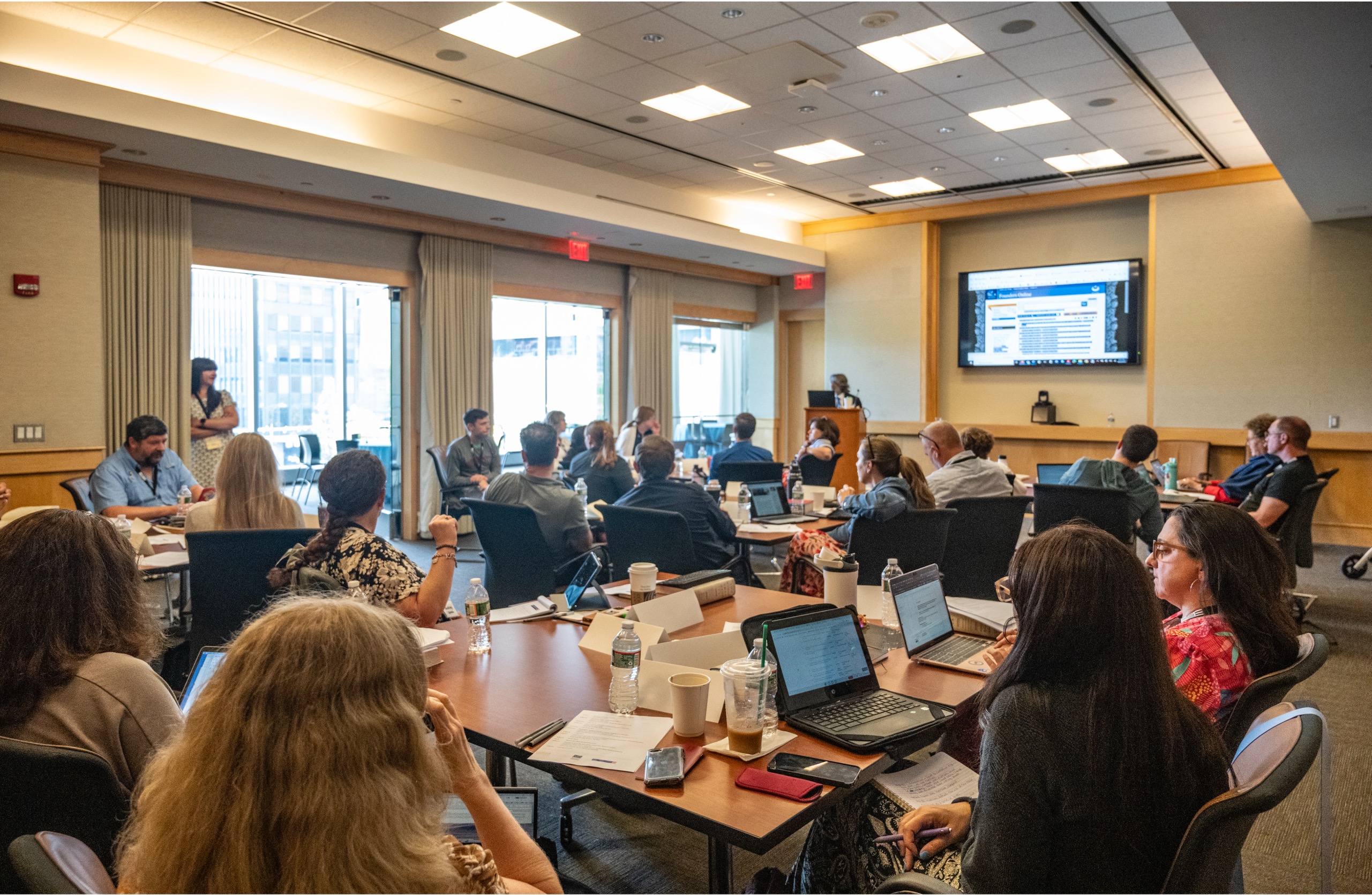 A group of people sit at tables in a conference room, watching a presentation on the Constitution displayed on a large screen. Some are using laptops, while others take notes or listen attentively.