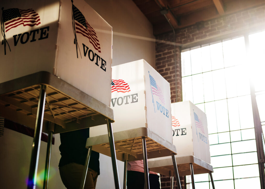 Four voting booths with American flags and "VOTE" signs are set up in a well-lit room, with sunlight streaming through large windows. People are partially visible casting their votes.